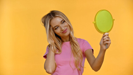Pretty blonde woman smiling while holding mirror isolated on yellow.