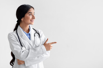 Smiling millennial indian woman doctor, family therapist in white coat show finger an empty space