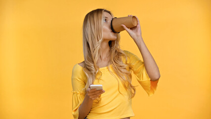 Blonde woman drinking coffee to go and holding smartphone isolated on yellow.