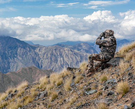 Shooter In Mountain Gear On The Slope Is Watching The Situation Through Binoculars.