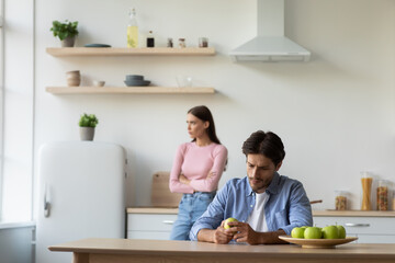 Obraz na płótnie Canvas Upset offended millennial caucasian husband ignores depressed unhappy wife after quarrel in white kitchen interior