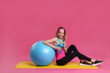 Fototapeta na wymiar Beautiful woman sitting on yoga mat with fitness ball against pink background