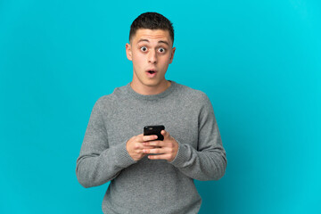 Young caucasian man isolated on blue background looking at the camera while using the mobile with surprised expression