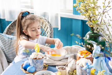 pretty little Armenian girl helps with baking for Easter on veranda on sunny spring day decorated with flowers and Easter decor, eggs, cake and willow branches, Easter family celebration