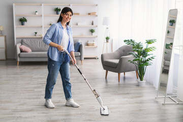 Cheerful woman cleaning floor with spray mop in living eoom