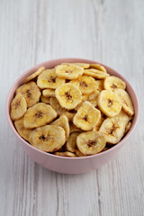 Homemade Banana Chips in a Pink Bowl on a white wooden surface, low angle view.