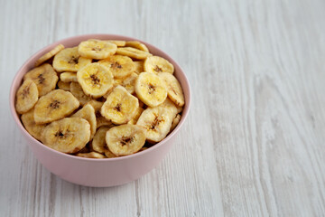 Homemade Banana Chips in a Pink Bowl on a white wooden background, side view. Space for text.