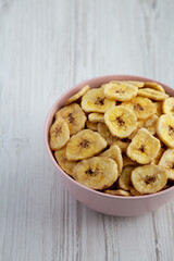 Homemade Banana Chips in a Pink Bowl on a white wooden background, side view.