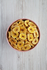 Homemade Banana Chips in a Pink Bowl on a white wooden background, top view. Flat lay, overhead, from above.