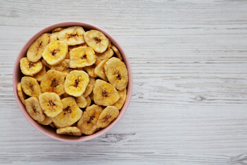 Homemade Banana Chips in a Pink Bowl on a white wooden background, top view. Flat lay, overhead, from above. Copy space.