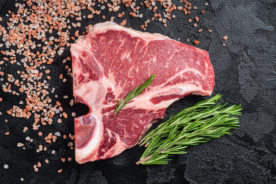 Raw T bone beef meat steak, porterhouse steak on butcher table with rosemary. Black background. Top view