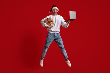 Fototapeta na wymiar Man In Santa Hat With Laptop And Gifts Jumping Over Red Background