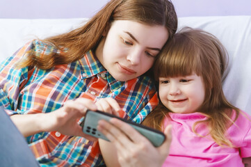 Happy sisters sitting on the couch watching cartoons together on a mobile phone. The older sister shows the cute younger sister a funny video on the smartphone screen online. Gadgets and kids.