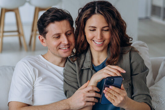 Young brunet-haired husband and curly wife discuss wedding photos from social media smiling broadly and sitting on sofa closeup