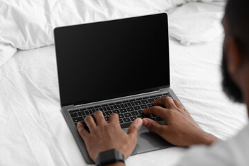 Young african american guy look at laptop with blank screen, work remotely on white bed in bedroom