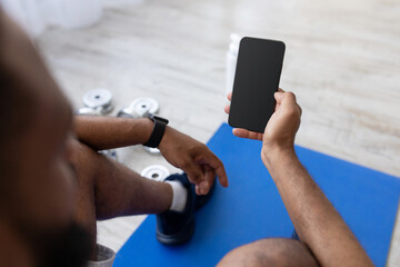 Millennial african american guy typing on smartphone with blank screen on mat on floor