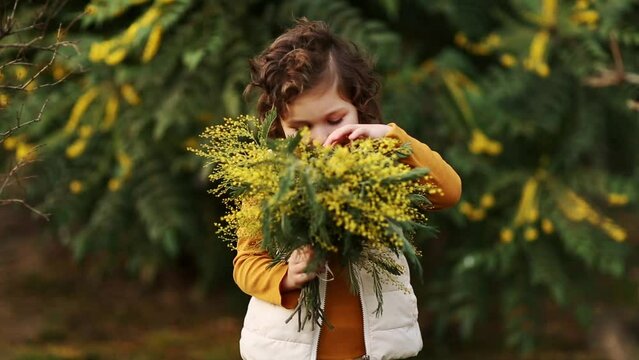 a small, beautiful girl is standing in the park in a yellow jacket and vest and holding a bouquet of yellow flowers in her hands
