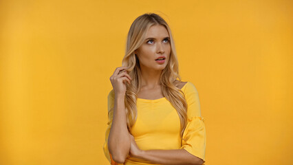 Pensive blonde woman touching hair isolated on yellow.