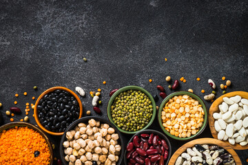Legumes, lentils, chikpea and beans assortment in different bowls on black stone table. Top view with copy space. Vegan food, protein source.