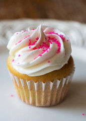 A close up view of a mini cupcake decorated with pink sugar sprinkles.  Confetti cupcake batter with white frosting on a white plate.  Wooden table blurred in background.