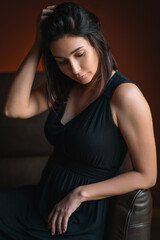 Naklejka premium Portrait of beautiful thoughtful woman titivating long dark hair in black dress sit reclining on brown leather sofa with eyes down. Depression, fatigue, stress, problems, emotions concept. Vertical. 