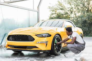 Car wash service outdoors. Car wash self-service concept. Handsome African guy wiping with green cloth car wheel during the washing process outdoors.