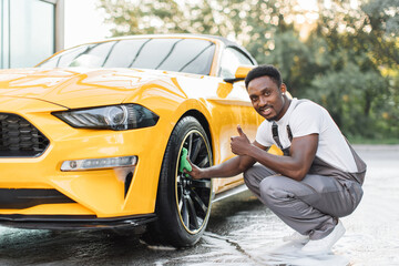 Car washing on open air. Young guy in gray overalls African bearded man cleaning a wheel while...