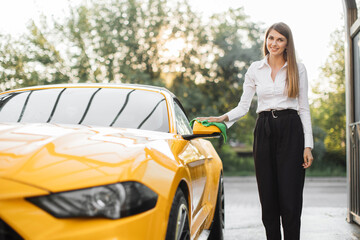Business woman wearing on a white shirt and black trousers wiping rearview car mirror of her modern luxury car in a self-service car wash station outdoors with a green microfiber cloth