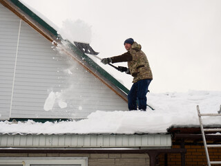 a man removes snow from the roof of a completely snow-covered house with a shovel. a lot of fresh...