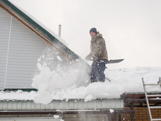 a man removes snow from the roof of a completely snow-covered house with a shovel. a lot of fresh snow after a blizzard, hard and dangerous work