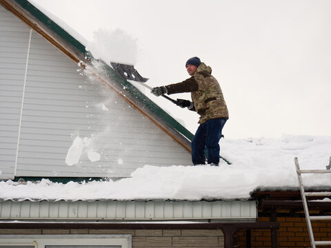 A Man Removes Snow From The Roof Of A Completely Snow-covered House With A Shovel. A Lot Of Fresh Snow After A Blizzard, Hard And Dangerous Work