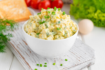 Egg salad with chopped green onions on top in a white bowl for cooking a sandwich on a white wooden...