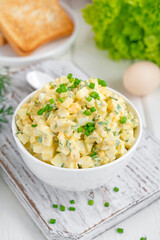 Egg salad with chopped green onions on top in a white bowl for cooking a sandwich on a white wooden background. Selective focus