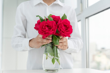 boy in white shirt puts red roses in big transparent vase, grreting and love concept, valentines day,