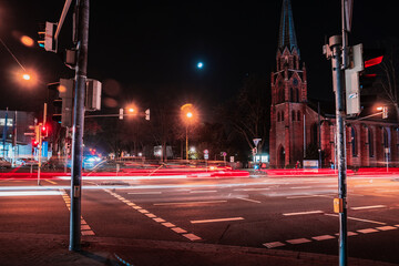 Light trails in night traffic on a Bremen rush hour street. Motion blur, red and blue lights of car, bus and cyclist. Red brick church in background. - obrazy, fototapety, plakaty