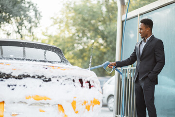Serious african man in business suit, at self car wash outdoors, is spraying cleaning foam to a...
