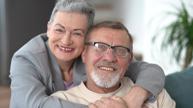 Man and a woman hugging and looking at the camera while sitting on the sofa. Indore portrait of a gray-haired elderly married couple
