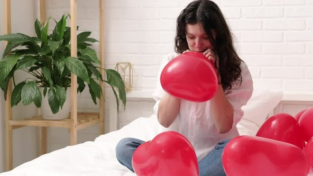 Girl is blowing air balloon at home Handsome brunette girl inflating red heart shaped balloon sitting on bed. Preparation for valentines day party, celebration, dating and surprise.