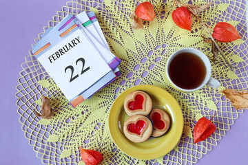 Calendar for February 22: the name of the month February in English, the numbers 22 on the loose-leaf calendar, a cup of tea, heart-shaped cookies, physalis branches on a yellow openwork napkin