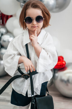 A fashionable baby in sunglasses is trying on her mom's red shoes. Fashion and modern clothes. Glitter bright metallic mirror background, new trends valentine's day