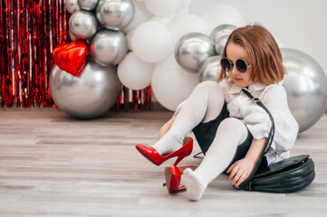 A fashionable baby in sunglasses is trying on her mom's red shoes. Fashion and modern clothes. Glitter bright metallic mirror background, new trends valentine's day