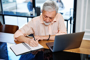 A happy senior student is sitting at home and writing down notes in a notebook while following an online lecture on the laptop. - obrazy, fototapety, plakaty