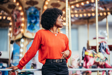 Obraz na płótnie Canvas Pensive young black woman with afro hairstyle and red sweater next to a carousel in the street