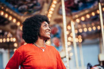 Close up of smiling dreamy young black woman with afro hairstyle and red sweater standing next to a carousel on the street