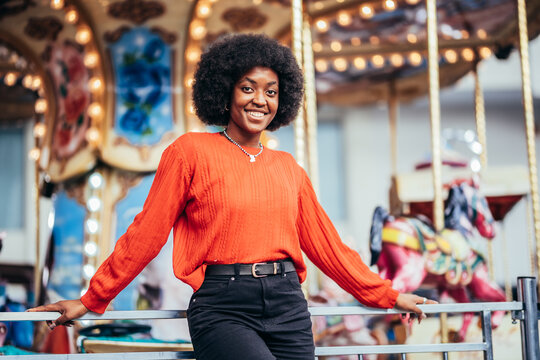 Smiling young black woman with afro hairstyle and red sweater next to a carousel on the street