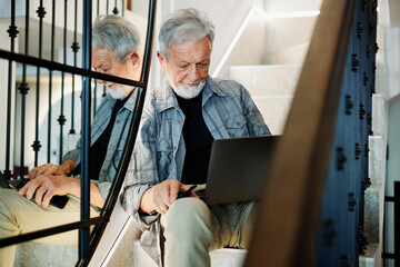 Fototapeta premium A happy senior student sits on the stairs at home with a laptop in his lap and using the it. He is checking on exam results online.