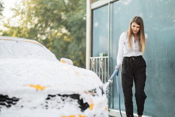 Attractive caucasian woman wear on business look washing his yellow car with foam at self service carwash outdoors. Clean car concept. Car wash.