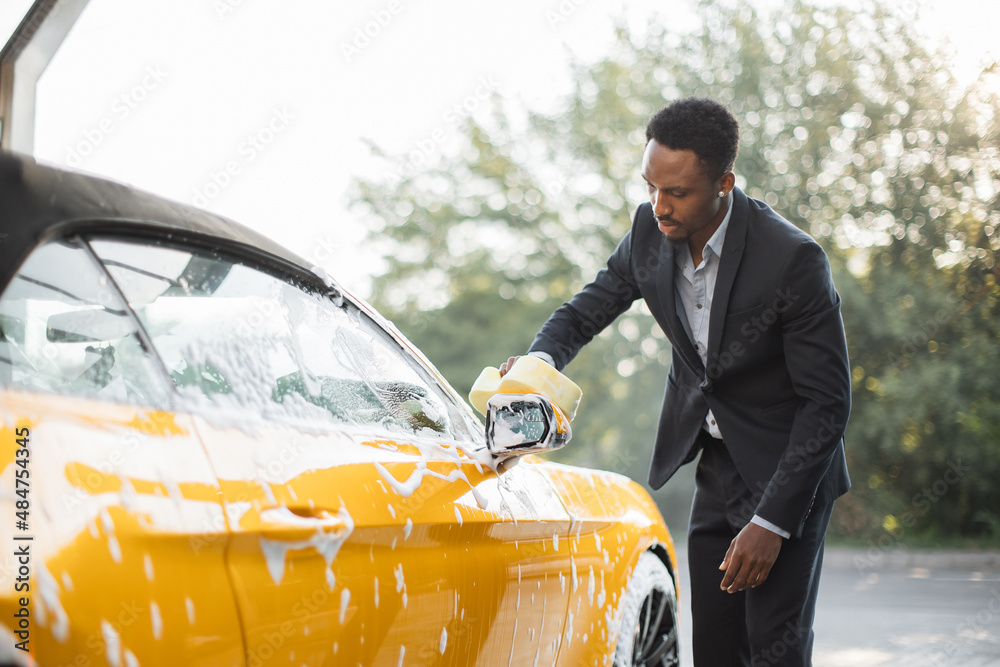 Sticker Handsome young African American businessman cleaning rearview car mirror with yellow sponge and soap foam outdoors at car wash service