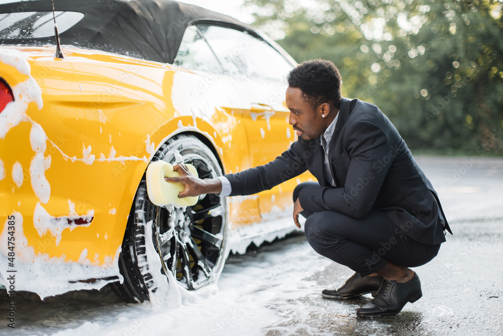 Wall mural Luxury modern sport car in soap foam outdoors at car wash service. Side view of handsome young African businessman using yellow sponge for cleaning rims outdoors.