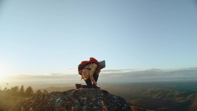 Female hiker kneel down on one knee to tie laces on hiking boot. Woman adventurer reaching summit on beautiful summer sunset. Authentic and cinematic wanderlust moment
